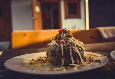 Ein Teller mit einer bunten, runden Speise, garniert mit Kräutern und Käse. Im Hintergrund sind Holzdetails und eine warme Atmosphäre zu erkennen.