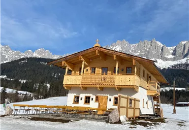 Ein schönes Holzhaus in den Bergen, umgeben von Schnee. Der klare Himmel und die majestätischen Berge im Hintergrund verleihen der Landschaft eine malerische Atmosphäre.