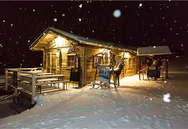 Eine gemütliche Berghütte im Schnee bei Nacht, mit festlicher Beleuchtung. Im Vordergrund sind Tische und eine Figur eines Rehs sichtbar.