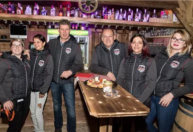 Eine Gruppe von sechs Personen steht in einer gemütlichen Bar mit Holzdekor. Sie tragen dunkelgraue Westen und lächeln freundlich in die Kamera.