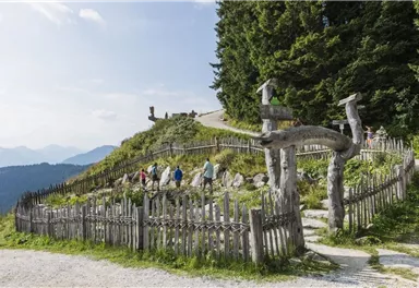 A picturesque mountain landscape with a wooden fence and several hikers. In the background, tall trees and the mountains are visible.