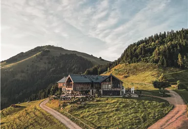 A picturesque mountain house in green landscape, surrounded by gentle hills and trees. The sky is blue with some clouds.