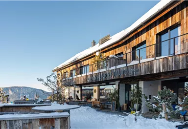 A modern wooden building in the snow with large windows and a clear blue sky. The surroundings are wintry and inviting.