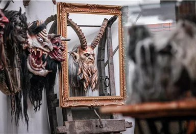 Ein Raum mit verschiedenen Masken an der Wand, darunter eine große, teufelsartige Maske. Im Hintergrund spiegelt sich eine weitere Maske in einem goldenen Rahmen.