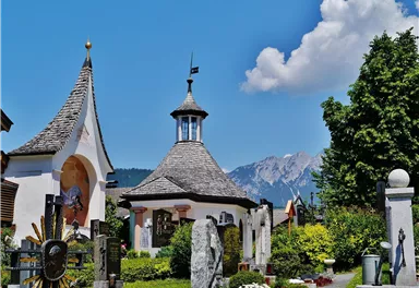 A picturesque place with a lovely chapel and green trees. In the background, impressive mountains can be seen under a blue sky.