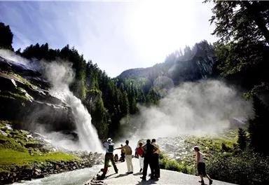 Eine Gruppe von Menschen steht in der Nähe eines beeindruckenden Wasserfalls umgeben von grünen Bäumen. Der Himmel ist klar und die Sonne strahlt hell.