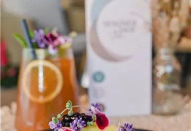 Ein köstlicher Kuchen mit einer Gericht aus frischen Kräutern und essbaren Blüten. Im Hintergrund sind zwei Getränke und eine Menükarte zu sehen.