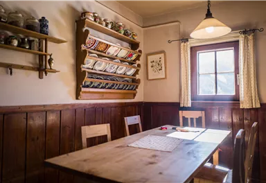 A cozy room with a wooden table and chairs. On the wall, dishes and storage containers are visible, while a window allows natural light to flood in.