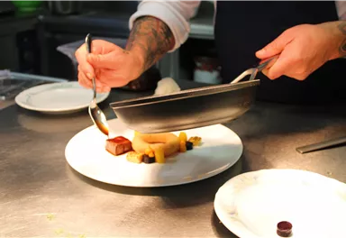 Ein Koch präsentiert ein kunstvolles Gericht auf einem weißen Teller. Verschiedene Zutaten werden sorgfältig angerichtet.