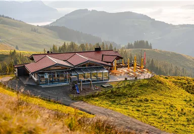 A modern building in the mountains with a terrace and wide green spaces. In the background, gentle hills and a clear sky can be seen.