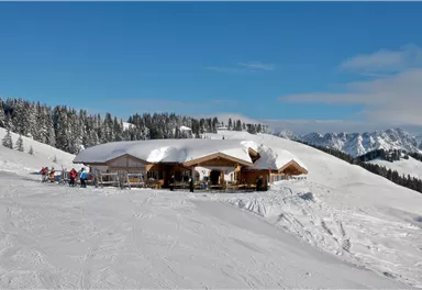 A cozy mountain cabin is located in a snow-covered winter landscape. In the background, there are snow-capped mountains and a clear blue sky.