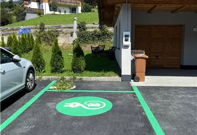 A parking lot with a green electric charging symbol. In the background, houses and trees can be seen.