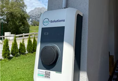 An electric charging station on a wall, surrounded by green areas and trees. The sky is blue with some clouds.
