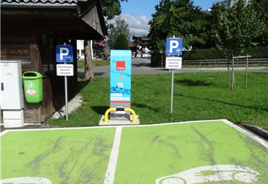 A parking lot for electric vehicles with a charging station and visible markings. In the background, there are additional parking spaces and a green area.