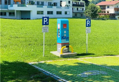 Ein Parkplatz mit einem Ticketautomaten steht auf einer grünen Wiese. Im Hintergrund sind Wohngebäude zu sehen.