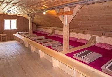 Ein einfaches Schlaflager mit Holzbetten und roten Bettdecken. Die Wände sind aus Holz, und es gibt ein Fenster, das Tageslicht hereinlässt.