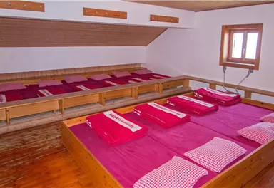 Ein schlichter Schlafraum mit mehreren Betten und roten Bettwäsche. Das Zimmer hat Holzverkleidung und Fenster, die Tageslicht hereinlassen.