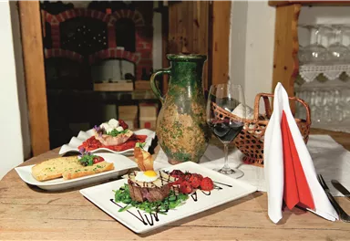 A beautifully set table with various dishes, including a steak on green leaves, a salad, and a piece of bread. There is also a wine glass and a decorative carafe.