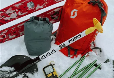A ski equipment lies in the snow, consisting of a red snowboard, an orange backpack, a shovel, and an avalanche probe. Additionally, other safety items and digital avalanche gear are visible.