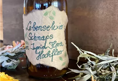 Eine Flasche mit beschriftetem Etikett für Schnaps steht auf einem Tisch. Um sie herum liegen getrocknete Kräuter und Früchte.