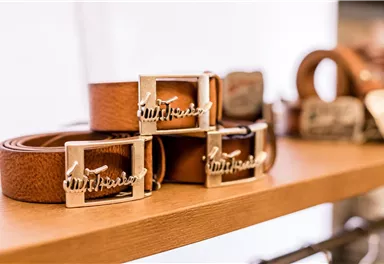 Drei Ledergürtel mit glänzenden Schnallen liegen auf einem Regal. Die Gürtel haben eine warme braune Farbe und sind stilvoll präsentiert.