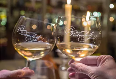 Two glasses of white wine are held up for a toast. In the background, a candle and a cozy atmosphere can be seen.