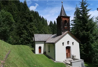 Eine kleine Kirche umgeben von dichten Bäumen. Der Himmel ist blau mit einigen Wolken.