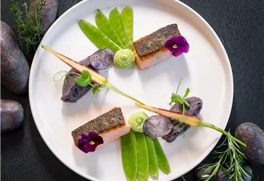 An elegant dish with grilled salmon, garnished with edible flowers and green peas. It is creatively presented on a white plate with purple potatoes in the background.
