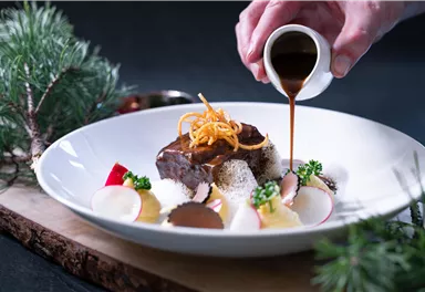 An elegantly arranged plate with meat and various side dishes. One person is pouring sauce over the dish.
