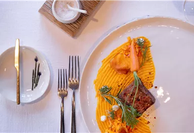 An artistically arranged plate with roasted meat, pureed pumpkin, and fresh herbs. Accompanied by matching cutlery and a small bowl of olives.