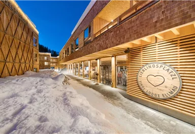 A modern building with wooden cladding, surrounded by snow. The entrance to the favorite place Lodge Ellmau is clearly visible, and the sky is clear and blue.