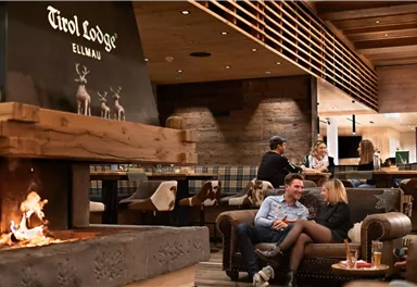 A cozy lodge with a large fireplace and wooden paneling. Couples relax in a warm atmosphere.