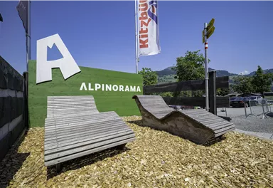 Ein Begrüßungsschild mit dem Schriftzug "Alpinorama" und zwei moderne Liegen aus Holz. Die Umgebung ist mit Rindenmulch dekoriert und zeigt eine klare blaue Himmel.