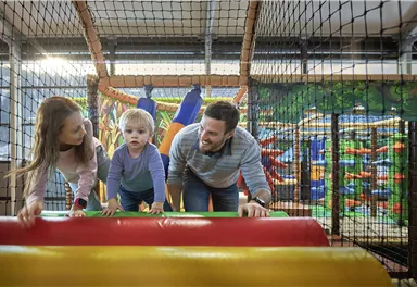 An indoor playground with colorful play equipment. A father is playing with his two children, who are climbing on a colorful obstacle course.