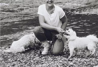 Eine fröhliche Frau kniet am Ufer eines Gewässers und hält zwei kleine Hunde an der Leine. Im Hintergrund sind Steine und Wasser sichtbar.