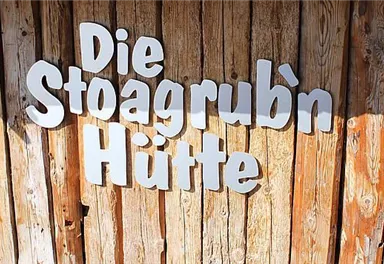 Eine Holzwand mit dem Schriftzug „Die Stoagrub’n Hütte“. Die Buchstaben sind weiß und hervorgehoben.