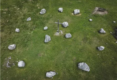 Eine grüne Wiese mit mehreren großen Steinen und einem Kreuz in der Mitte. Im Hintergrund sind einige weitere Steine und eine kleine Sitzgelegenheit sichtbar.