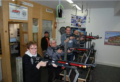 A group of five people is standing in a room holding sports rifles. They appear to be participating in a shooting sports event.