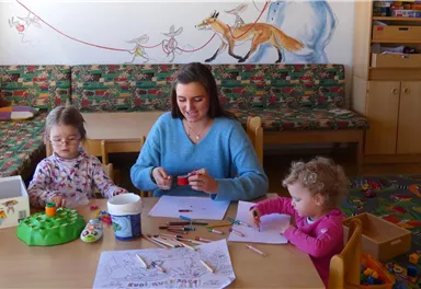 Eine Frau sitzt an einem Tisch mit zwei kleinen Mädchen und bastelt. Um sie herum sind verschiedene Bastelmaterialien und ein freundliches, buntes Zimmer.