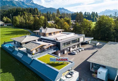 A modern building in a rural setting, surrounded by trees and mountains. There are parking spaces and a large meadow in the foreground.