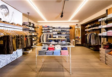 Ein modernes Bekleidungsgeschäft mit ordentlich angeordneten Regalen und einer Vielzahl von Kleidung. In der Mitte steht ein Glastisch mit gefalteten Oberteilen.