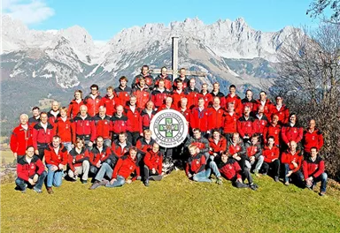 A large group of people in red jackets stands on a meadow in front of majestic mountains. They are posing around a shared sign in the middle of the picture.