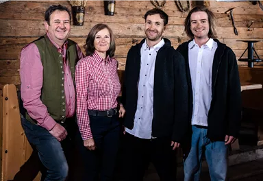 Eine Gruppe von vier Personen steht lächelnd vor einer rustikalen Holzwand. Sie tragen legere, moderne Kleidung und wirken freundlich und einladend.