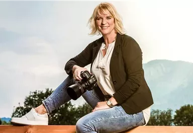 A smiling woman is sitting on a wooden bench, holding a camera in her hand. Trees and mountains are visible in the background.