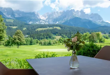 A beautiful table with a vase of flowers stands in front of a picturesque mountain landscape. In the background, green meadows and tall mountains can be seen.