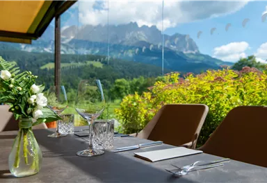 An elegant table with a view of mountains and a green landscape. The table is decorated with glasses and flowers, creating an inviting atmosphere.