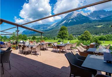 A terrace with tables and chairs offers a view of impressive mountains and a green landscape. The sky is clear with some clouds.