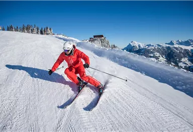 Ein Skifahrer in roter Kleidung fährt über den Schnee in den Bergen. Der klare blaue Himmel und die verschneiten Gipfel im Hintergrund schaffen eine wunderschöne Winterlandschaft.