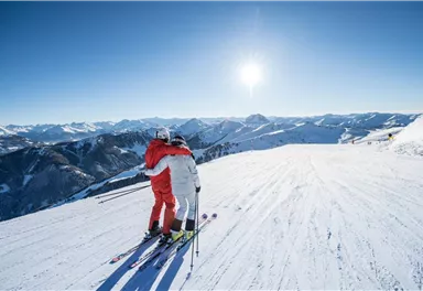 A couple stands on a snow-covered slope and embraces each other. In the background, the mountains can be seen under a clear blue sky.