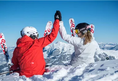 A couple with ski equipment sits in the snow and gives each other a high-five. In the background, snow-covered mountains and a blue sky can be seen.
